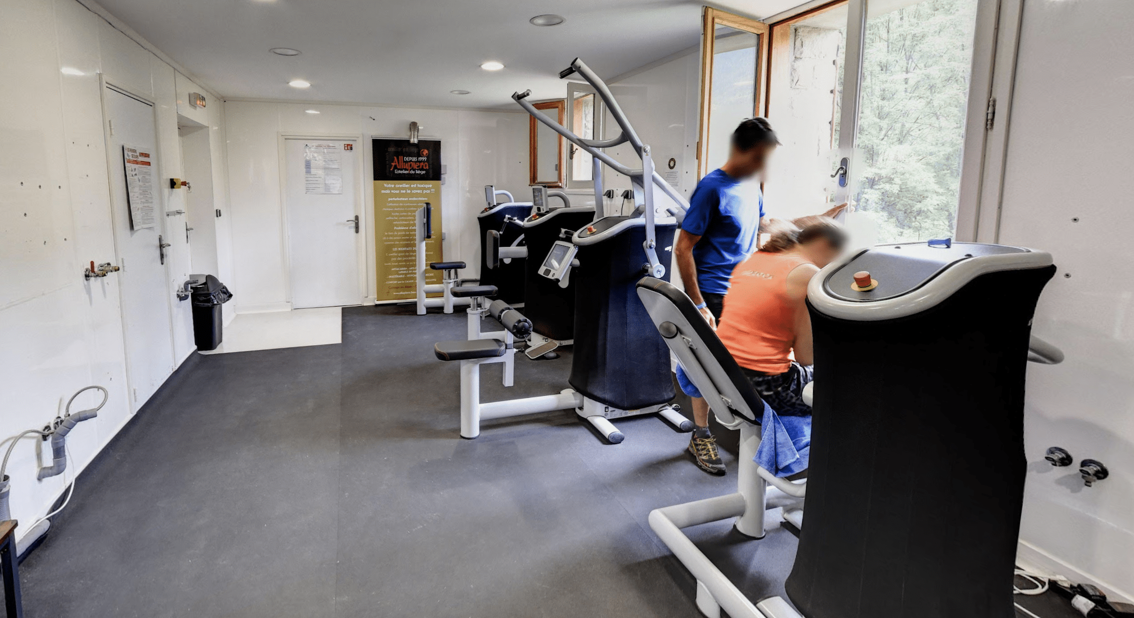 Intérieur d'une salle de sport avec des appareils de musculation. Des personnes s'entrainent près d'une fenêtre ouverte. Panneau d'informations Allupiera.