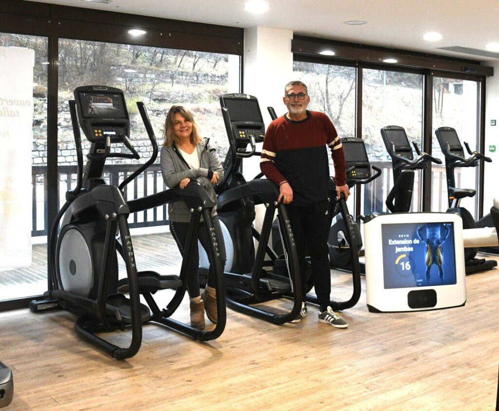 Couple souriant dans une salle de sport, posant à côté de vélos elliptiques Matrix. Écran affichant un exercice de jambe. Fitness, santé.
