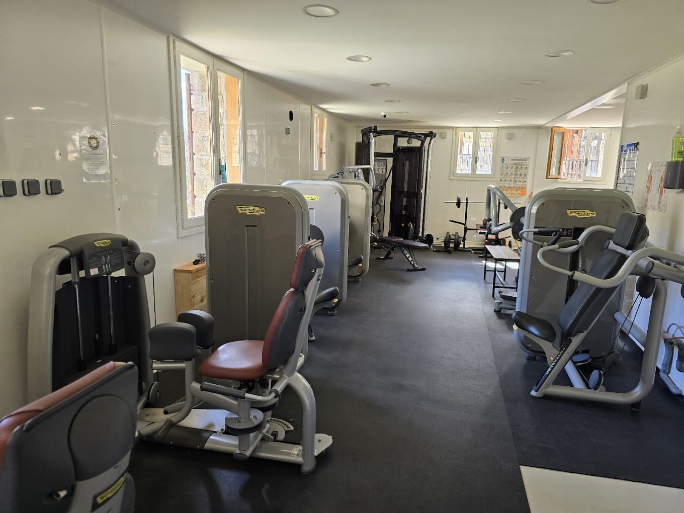 Intérieur de salle de sport lumineuse avec machines Technogym, haltères et fenêtres. Ambiance fitness et bien-être dans un espace moderne.