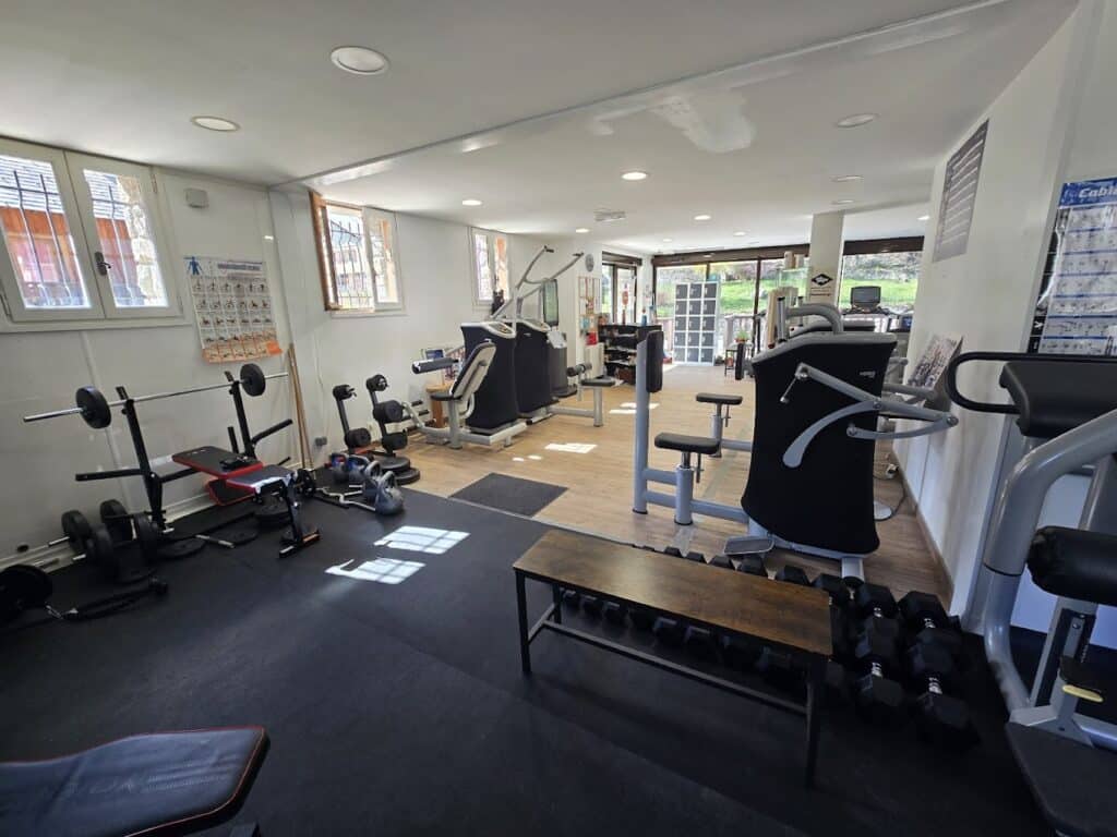 Salle de sport lumineuse avec équipements de musculation variés : haltères, banc, machines guidées. Affiche d'exercices visible. Vue sur la nature depuis les fenêtres.