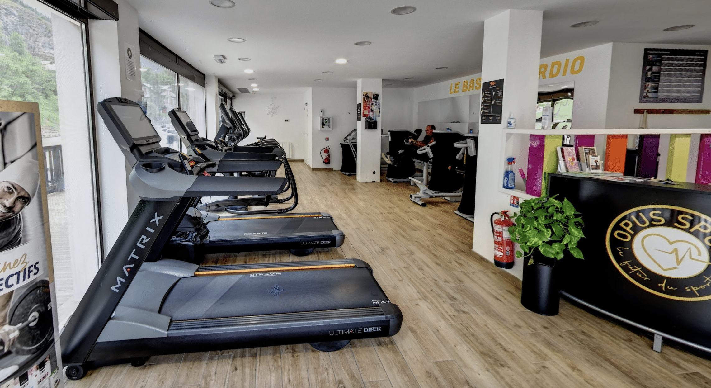 Salle de sport lumineuse avec tapis de course Matrix Ultimate Deck, zone cardio. Affiche, plante, et comptoir d'accueil Opus Sport visibles.