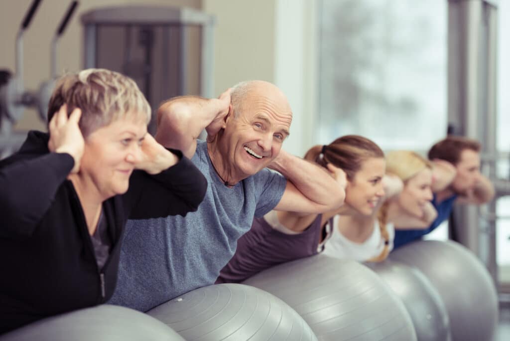 Groupe varié (seniors, jeunes) fait des abdos sur des ballons d'exercice en salle de sport. L'homme senior sourit.